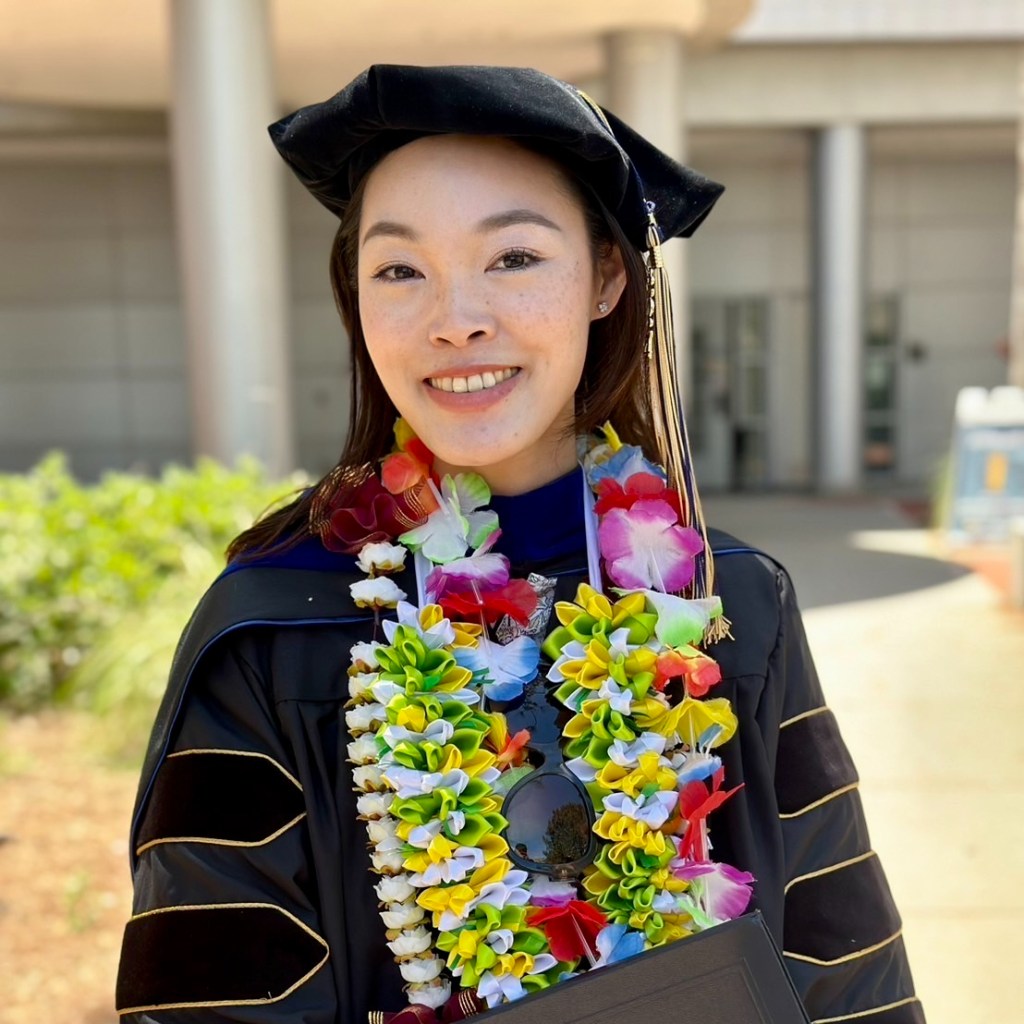 Doreen Hsu, Ph.D. is shown smiling and wearing her doctoral graduation regalia. She is standing in front of an academic building.