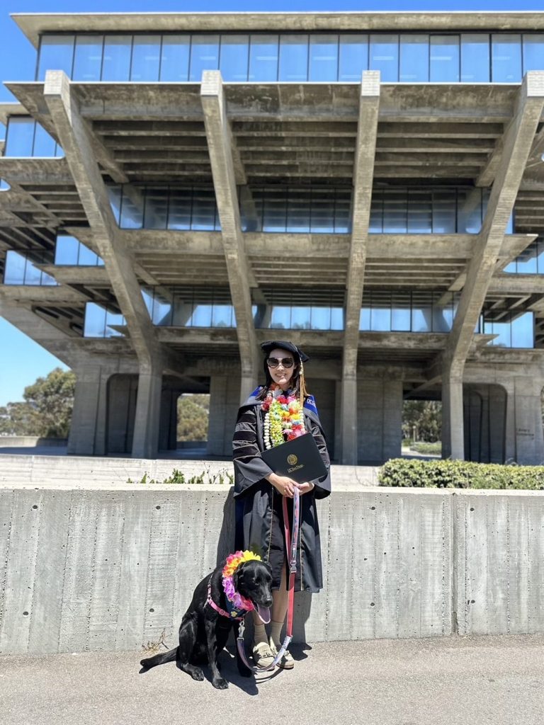 Tiffany the black labrador dog and myself, Doreen, wearing our Ph.D. graduation regalia at the UCSD Geisel Library. Doreen is wearing the black doctoral gown, cap, tassel, and royal blue mantle sash. Tiffany is wearing a lei flower necklace. Doreen and Tiffany are standing on a walk path in front of the Geisel Library structure, which features glass window panes that reflect the sunny blue sky.