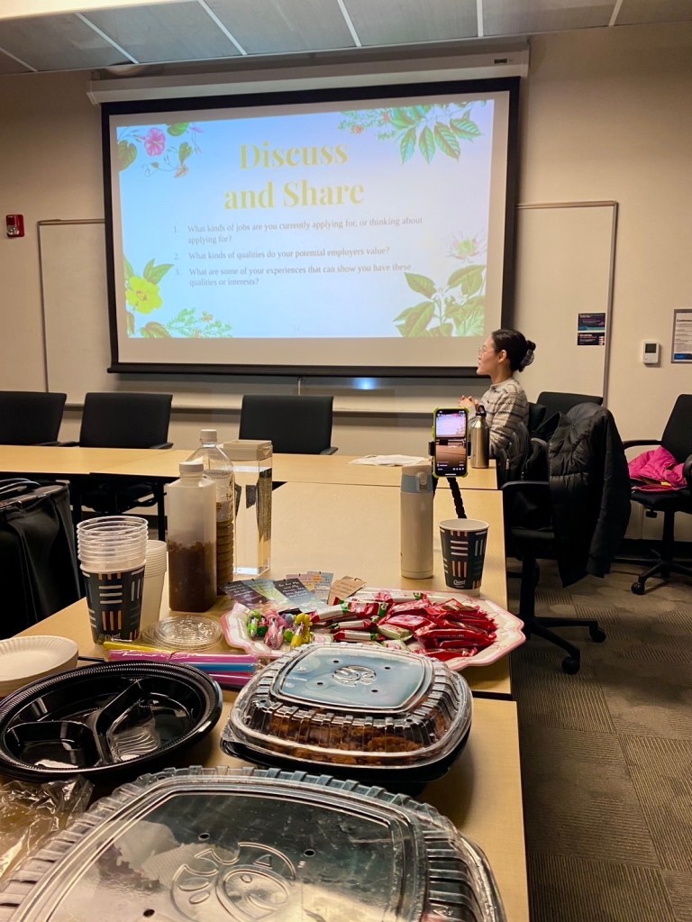 Image of Doreen Hsu, presenter, at the Cover Letters and Resumes Workshop. Doreen is sharing some discussion questions shown on the projector screen. In the foreground is a spread of food, drinks, snacks, and special gifts for audience members. 