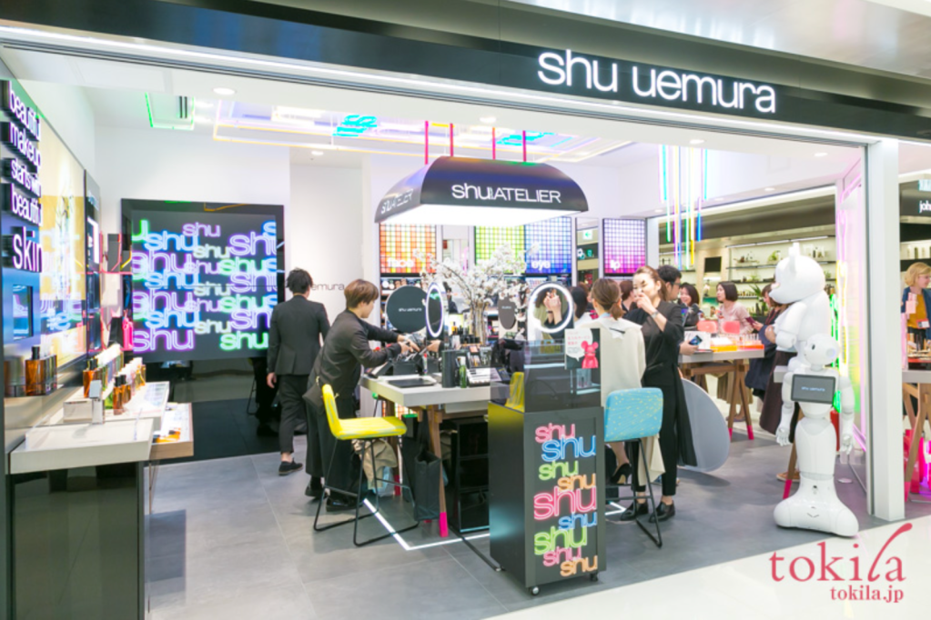 Image of shu uemura brand makeup and skincare store. There are several staff and customers inside the store space, in where there is a large table and seats in the center for makeup demos. 