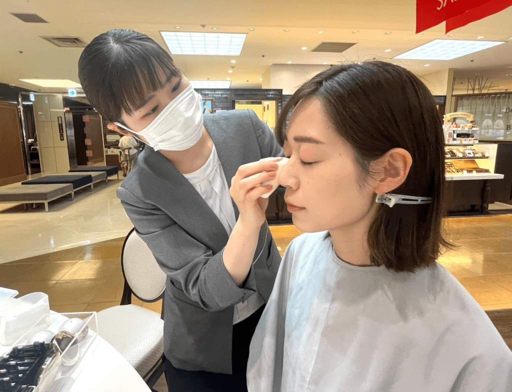 Image of a beauty advisor applying a makeup product onto a customer, who is wearing an apron and hair clips to keep her hair out of the way of product application. 